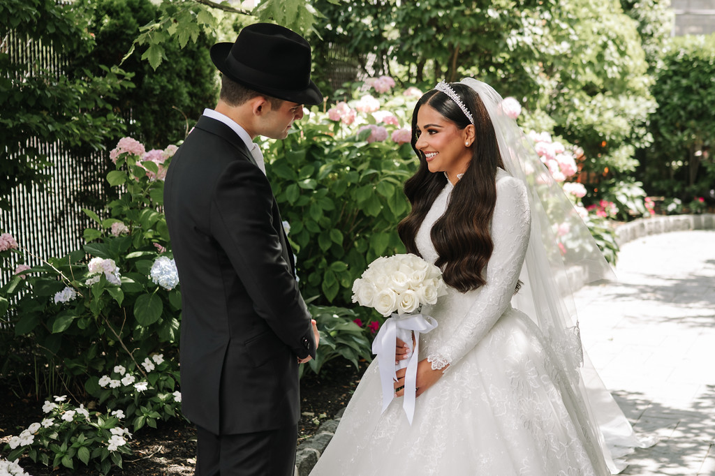 Outdoor wedding portrait in a garden: groom in a black hat and suit faces the smiling bride in a white lace gown and veil, holding a bouquet of white roses.