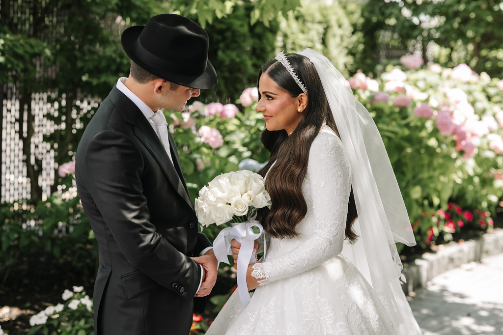Frum wedding couple standing face-to-face outdoors; the groom leans in while the bride smiles in her wedding dress holding a white bouquet and veil.
