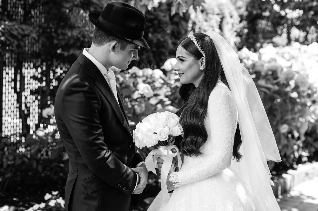 Black-and-white wedding portrait of a bride and groom facing each other, smiling as they hold a white bouquet outdoors under trees.
