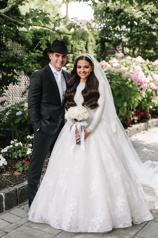 Frum newlywed couple posing outdoors in a garden path, the groom in a black hat and suit and the bride in a white lace bridal gown and veil, smiling while holding a bouquet.