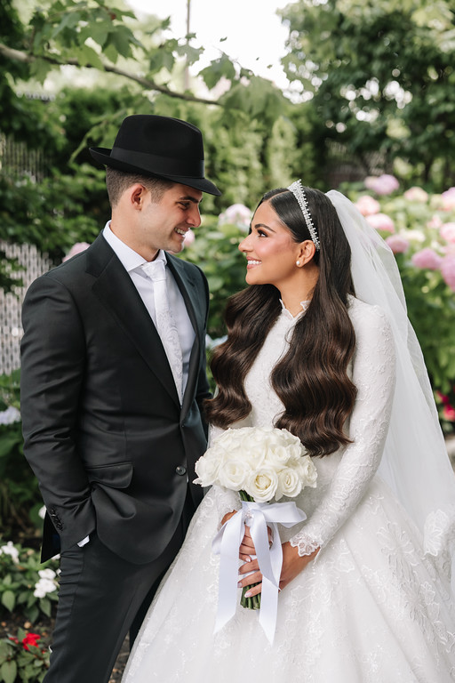 Frum wedding portrait of a groom in a black hat and suit and a bride in a white lace gown with veil, smiling while holding a bouquet in a garden.