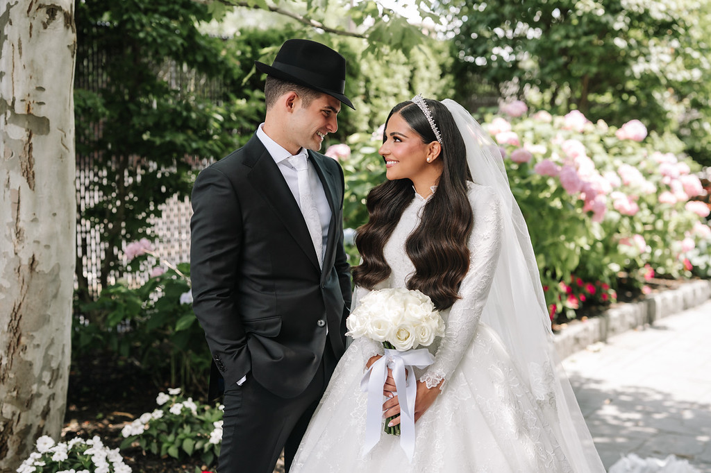 Bride in a white gown and veil holds a bouquet while smiling at her groom in a black hat and suit during a close moment outdoors.
