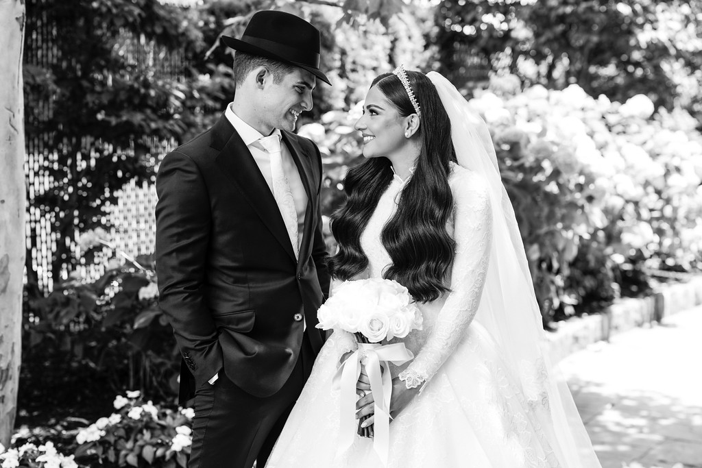 Black-and-white outdoor portrait of a Jewish bride and groom standing close, smiling at each other as the bride holds a bouquet.