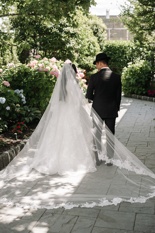 Bride and groom holding hands in a sunny garden path, surrounded by pink and white flowers, seen from behind as they walk toward the chuppah area.