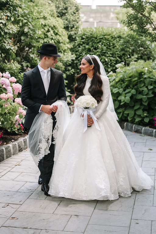Wedding couple holding hands and smiling outdoors in a garden walkway, with the bride in a white gown and veil and the groom in a black hat and suit.