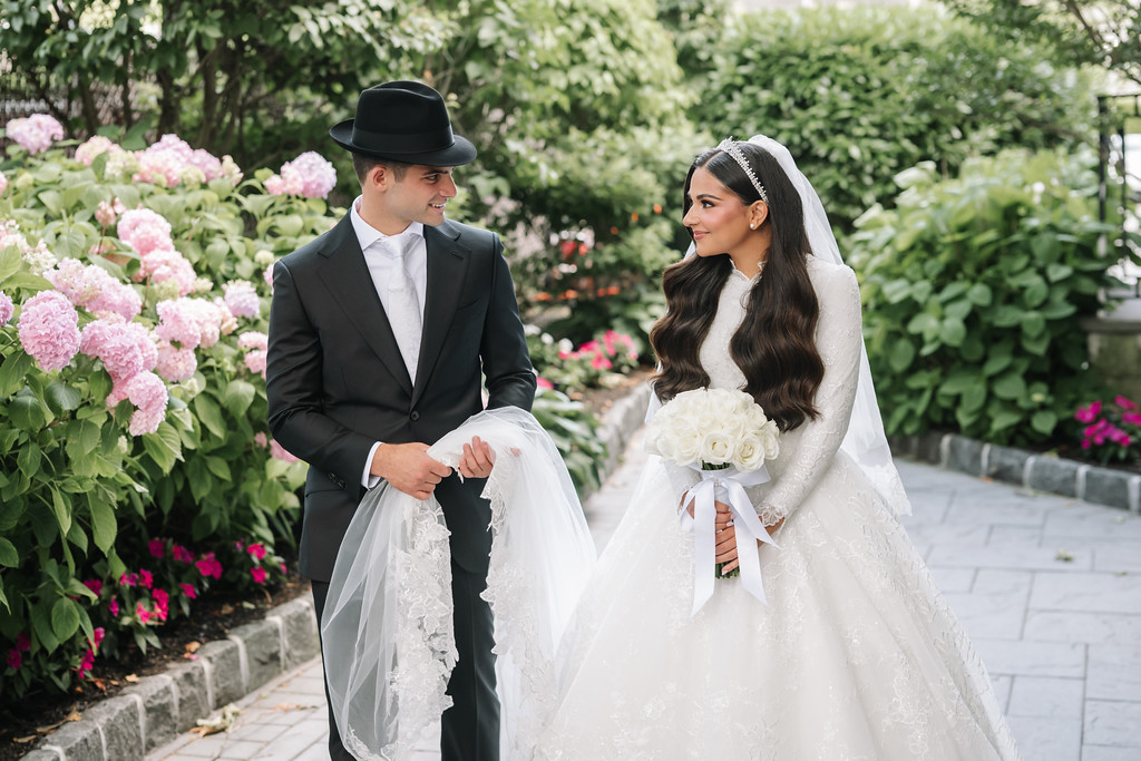 Outdoor wedding portrait of a couple walking together in a garden, the groom in a black hat and suit and the bride in a white lace gown and veil, holding a bouquet, smiling at each other.