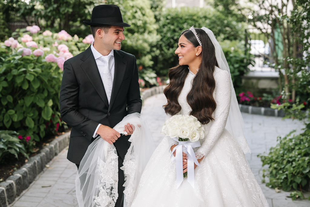 A bride in a white dress and veil smiles beside her groom in a black hat and suit as they hold hands on a garden walkway.