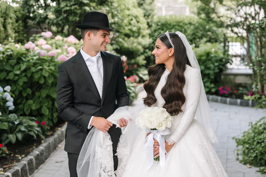 Frum wedding couple in formalwear—groom in black hat and suit and bride in a white lace gown with veil—holding hands and smiling at each other outdoors among flowers.