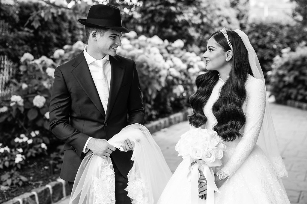 Black-and-white portrait of a groom in a black hat and suit smiling at his bride in a white wedding dress and veil, standing outdoors among flowers.
