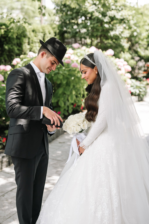Couple during badeken outdoors: the chosson in a black hat leans in while the kallah in a white bridal gown and veil holds her bouquet, looking at him.