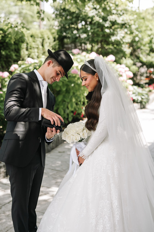 Frum wedding badeken moment outdoors: groom in a black suit and black hat gently adjusts his hands toward the bride in a white gown and veil; they smile while facing each other.