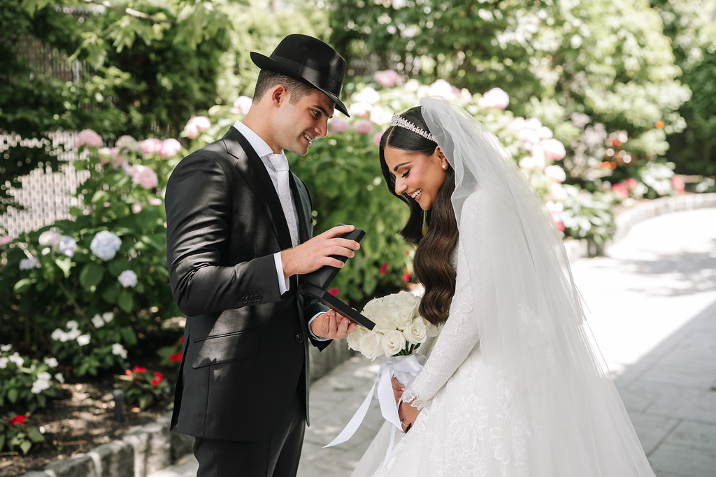 Bride in a white veil and groom in a black hat during badeken, smiling at each other in a garden ceremony setting.