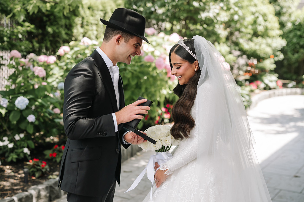 A groom in a black hat and suit and a bride in a white wedding dress and veil smile at each other during the badeken, holding a small bouquet with lush garden flowers in the background.