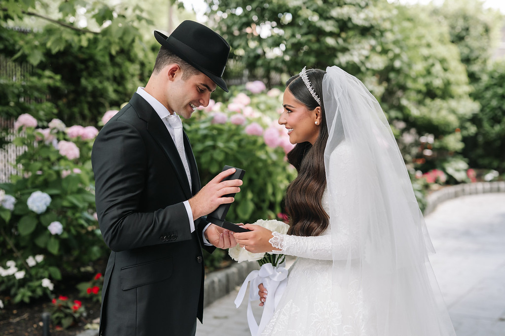 Frum wedding couple during badeken outdoors: the groom in a black hat and suit smiles while opening a small item as the bride in a white dress and veil looks at him in a garden setting.