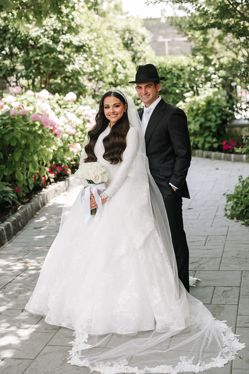 Smiling bride in a white lace gown and veil and groom in a black hat and suit posing together on a garden walkway surrounded by blooming flowers.