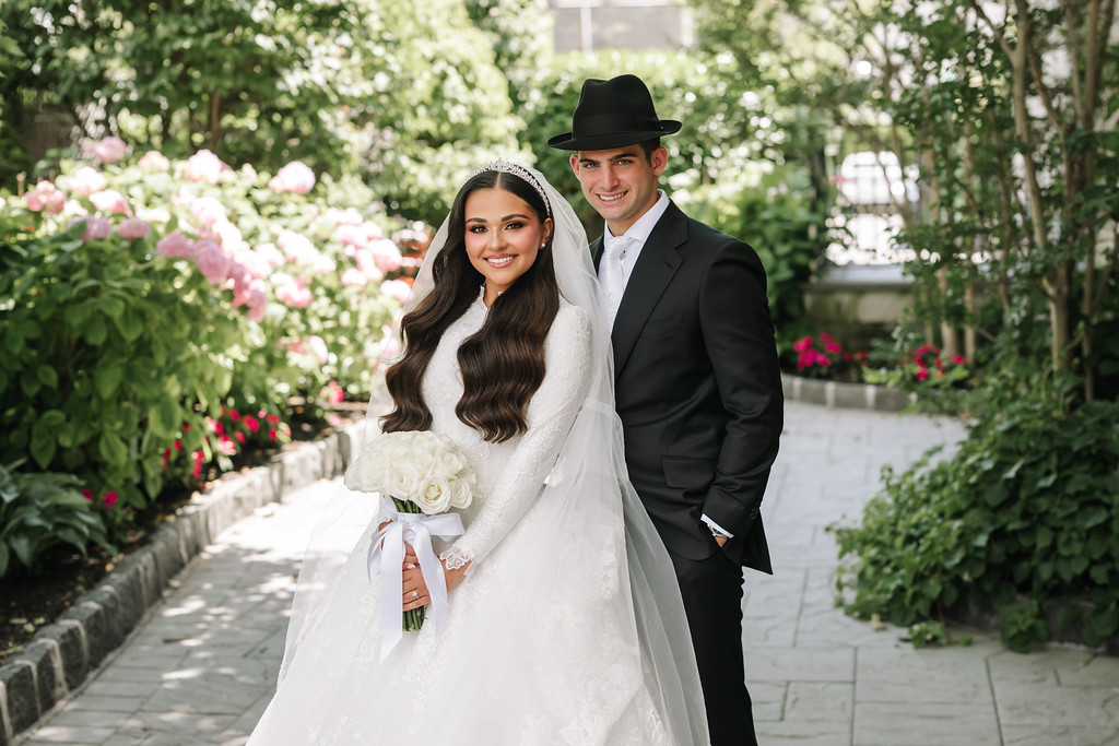 Frum bride and groom in wedding attire standing on a garden walkway, smiling with the bride holding a bouquet as they pose outdoors.