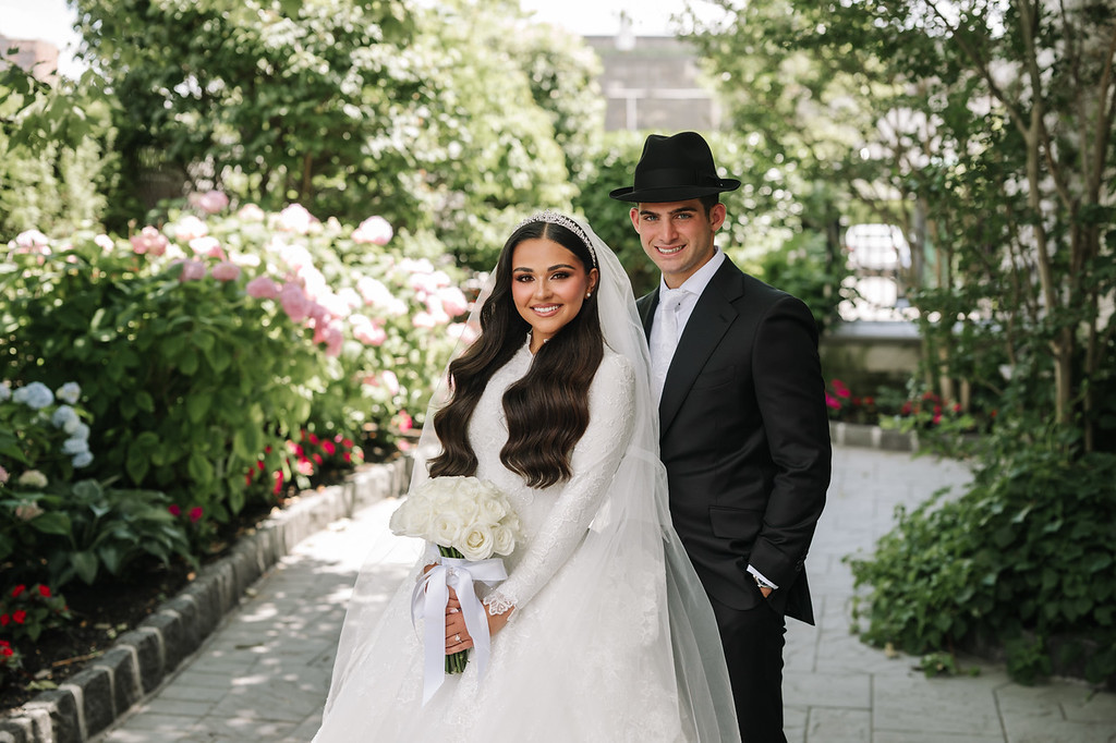 Smiling bride and groom in formal wedding attire standing together in a lush garden walkway with pink flowers and greenery behind them.