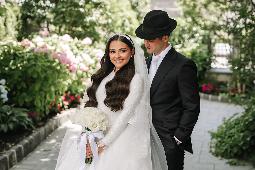 Frum wedding badeken outdoors in a garden: the bride in a white dress and veil smiles while holding a bouquet, with the groom in a black hat standing beside her.