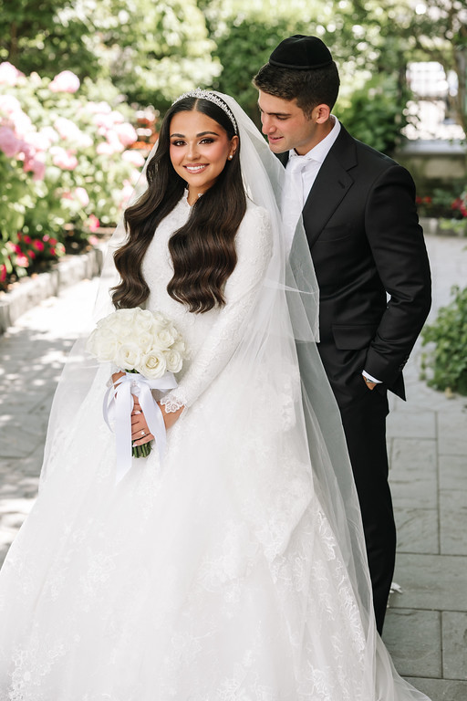Smiling bride in a white lace gown and veil holding a bouquet, with the groom beside her in black attire during a wedding portrait outside.