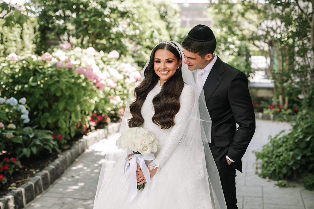Frum bride in a white bridal gown and lace veil smiles while holding a bouquet as the groom stands close beside her on a garden path lined with flowers.