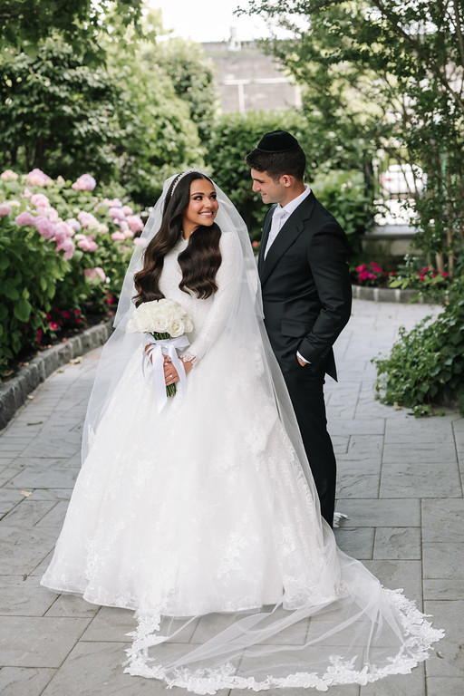 A bride in a white wedding gown and veil holds a bouquet as she smiles outdoors on a wedding day walkway, with the groom standing beside her in a black kippah and suit.