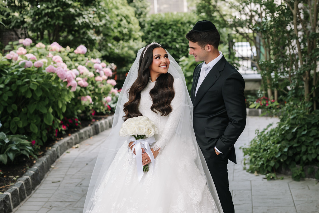 Frum bride in a white lace gown and veil smiling while holding a bouquet outdoors, with her groom in a black suit and kippah looking at her beside her in a garden walkway.