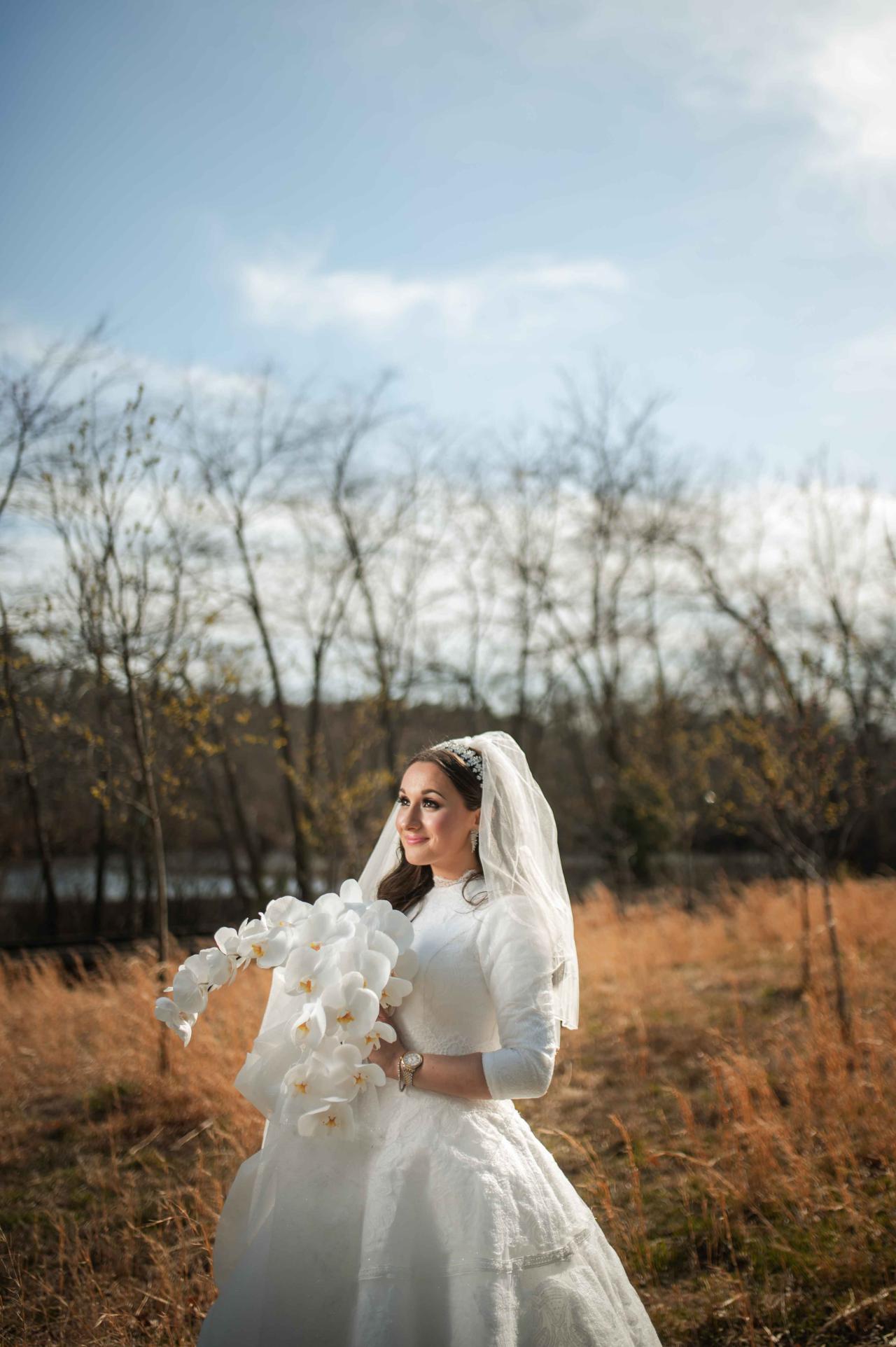 Frum bride in a white wedding dress and veil holding a bouquet of white orchids outdoors at golden hour, looking at the camera.