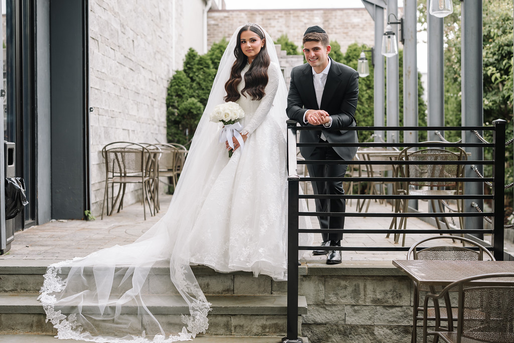 Frum bride and groom posed for a wedding portrait, standing side-by-side on steps outdoors with the bride in a white lace gown and veil and the groom in a dark suit.