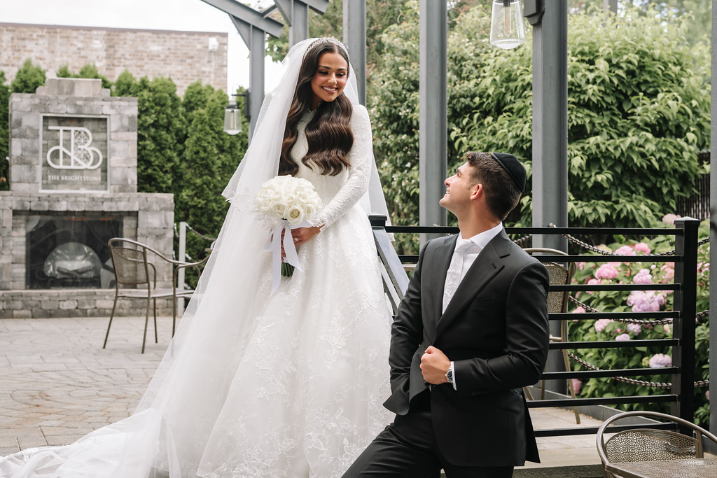 A bride in a white wedding gown and veil smiles while holding a bouquet near the chuppah as the groom stands beside her holding her hand.