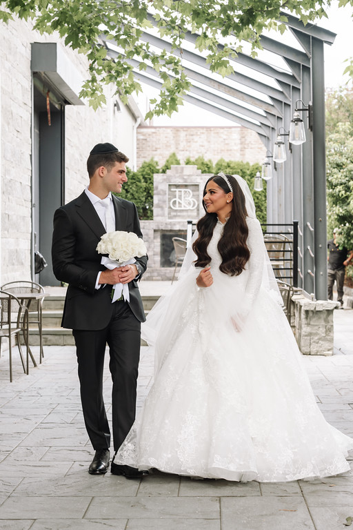 Frum newlyweds in wedding attire smiling at each other while walking outdoors in a courtyard, bride’s veil and dress flowing.