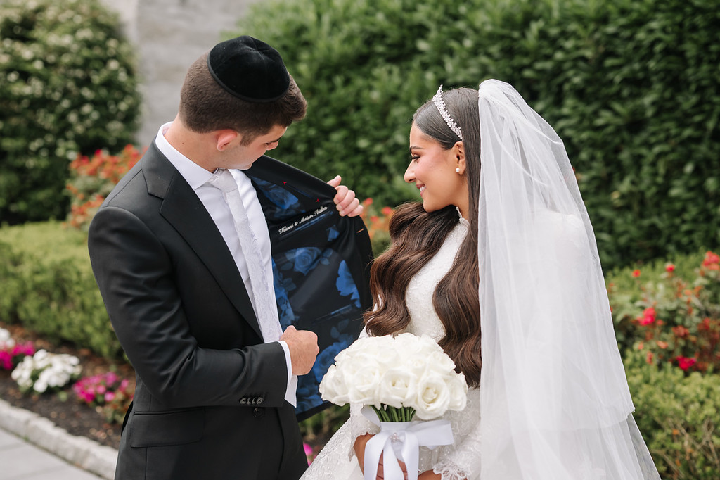 Frum wedding couple during badeken outdoors: groom in a black suit and kippah holds the bride’s hand while she smiles in her white veil and wedding dress.