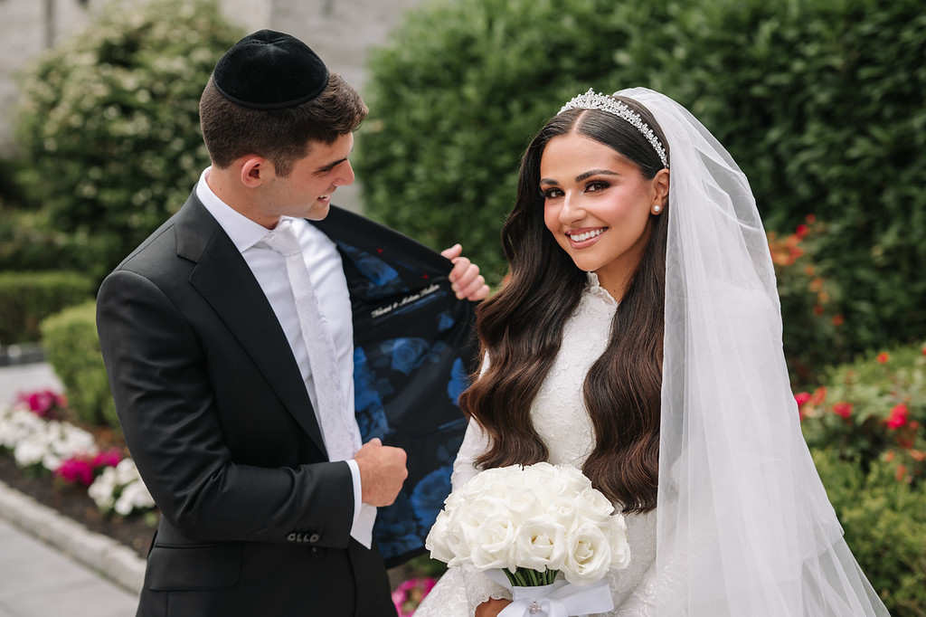 Smiling bride in a white veil holding a bouquet as the groom in a black kippah and suit looks at her during an outdoor wedding moment.