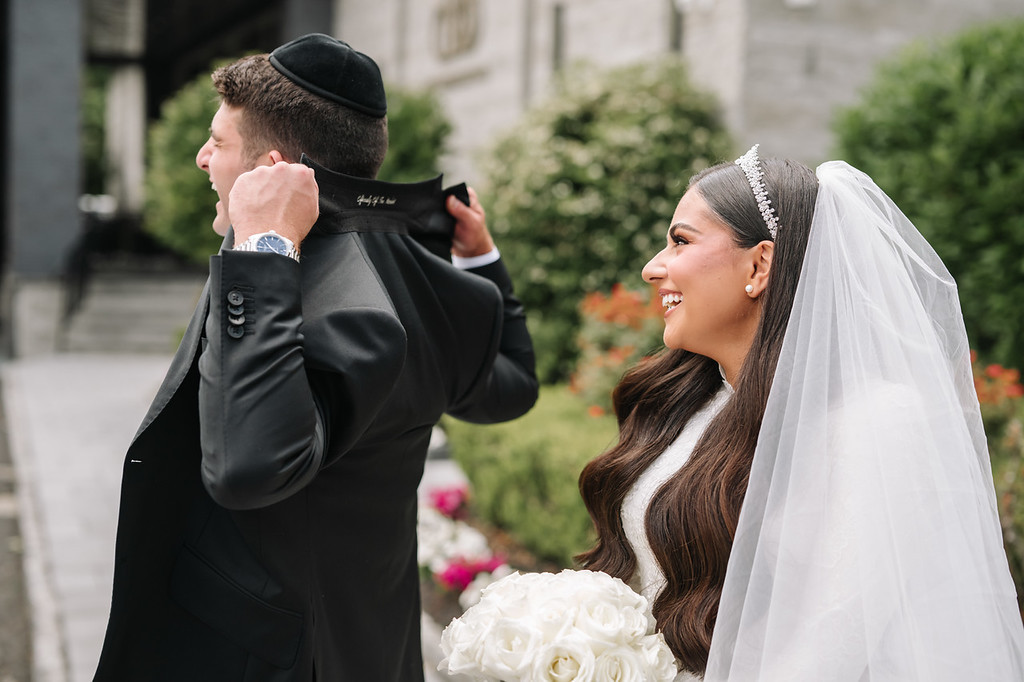 Smiling bride in a white wedding veil and headpiece laughs as the groom, in a black kippah and suit, gently adjusts her veil outdoors near the ceremony space.