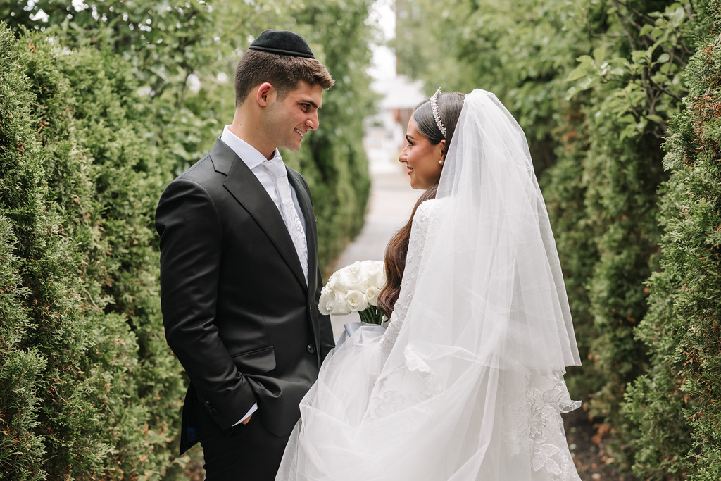 Bride in a white wedding veil and groom in a dark suit stand facing each other, smiling closely with greenery-lined hedges in the background.