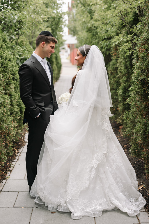 Bride and groom standing close in a garden walkway, smiling while the bride’s wedding veil covers part of her head and shoulders during the moment before the ceremony.