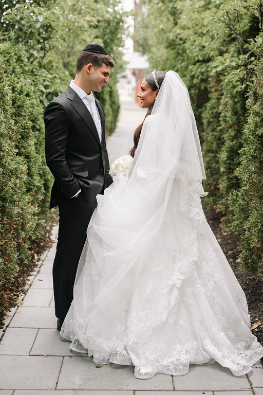 Bride and groom stand facing each other on a garden walkway, smiling warmly during their wedding ceremony moment.