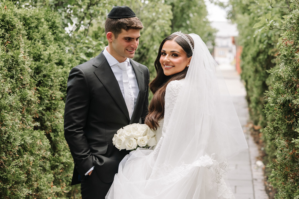 Bride in a white lace gown and veil smiles next to the groom in a black suit and black kippah as they pose along a green garden walkway.