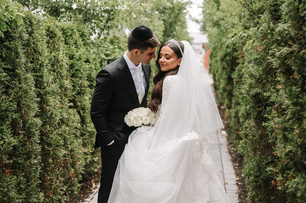 Bride in a white lace veil and groom in a black suit stand close together on a garden walkway, smiling as they hold flowers during the badeken moment.