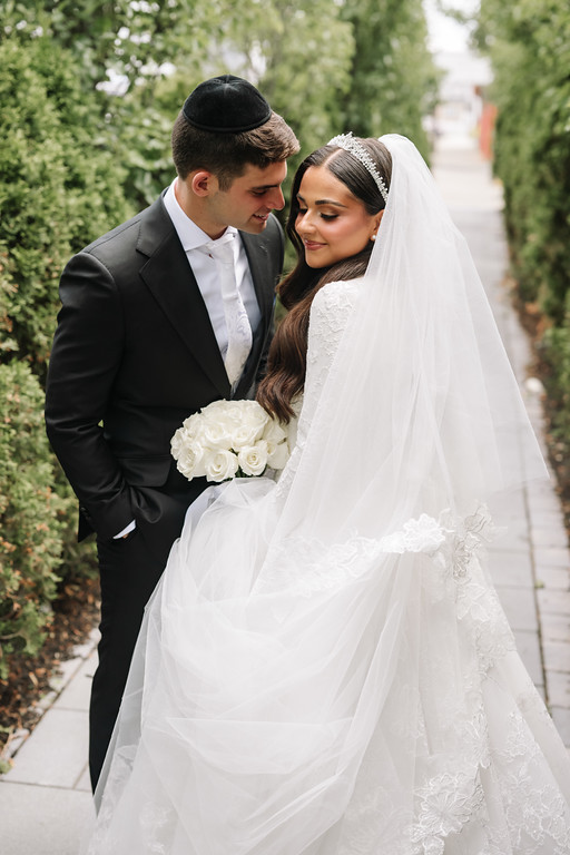 Frum wedding couple standing close outdoors on a walkway, bride in a white lace headpiece and veil holding a bouquet while the groom leans in and smiles at her.