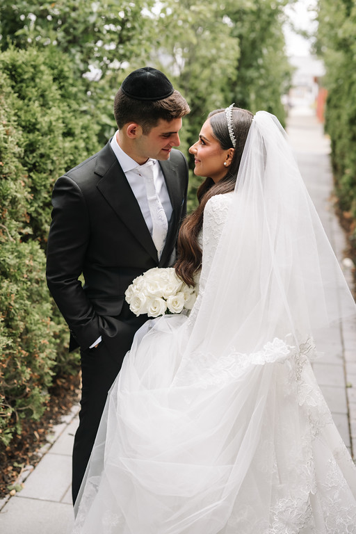 A groom in a black suit and kippah leans in toward his bride in a white lace wedding dress and veil as they smile and embrace outdoors with greenery in the background.