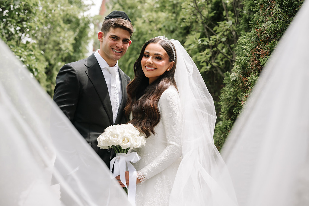 Outdoor wedding portrait of a smiling bride in a white veil and gown holding a bouquet, with the groom standing beside her among green trees.