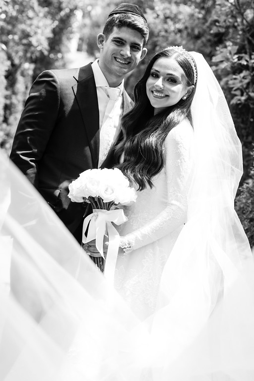 Black-and-white outdoor portrait of a bride and groom smiling closely together, the bride in a lace gown and veil holding a bouquet, with greenery in the background.