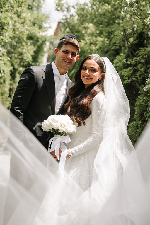 Newlywed couple standing close outdoors, smiling with wedding veil and suit, bouquet in the bride’s hands.