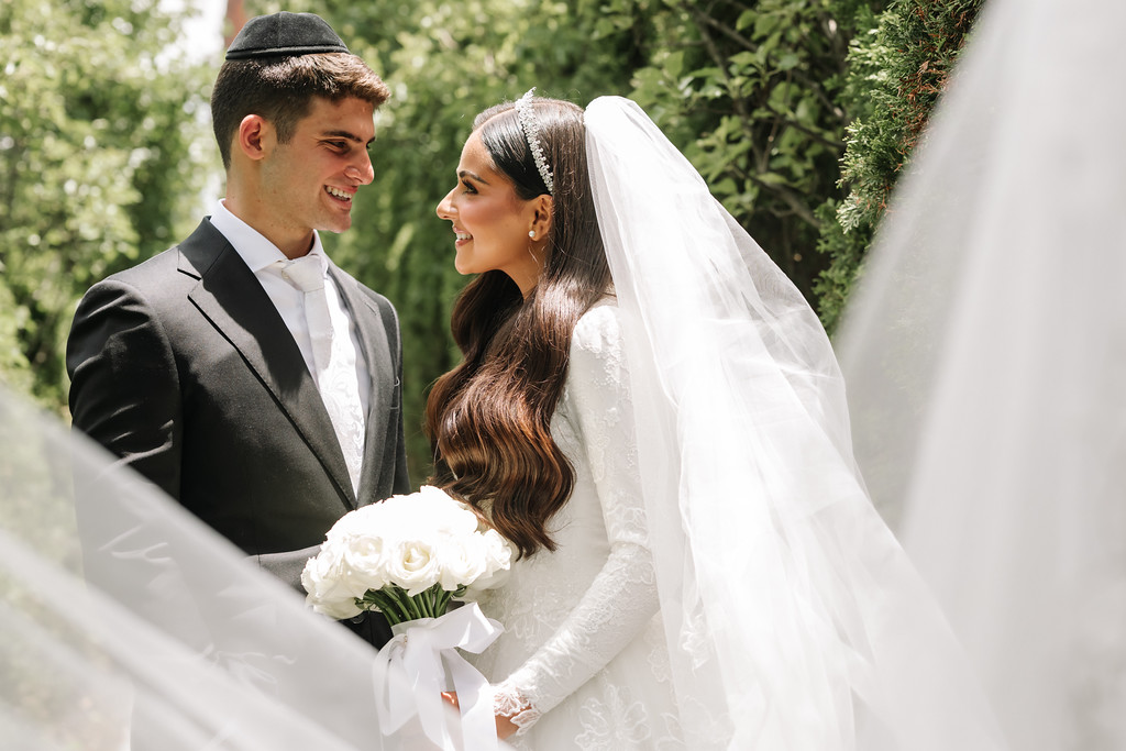 Outdoor wedding photo of the bride and groom smiling and holding hands, the bride’s veil in the foreground, with lush greenery in the background.