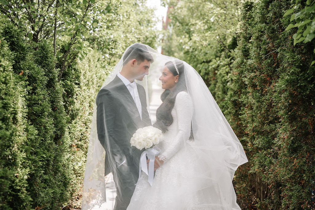 A groom and bride in wedding attire share an intimate moment during the badeken, standing among lush greenery with the bridal veil softly covering them.