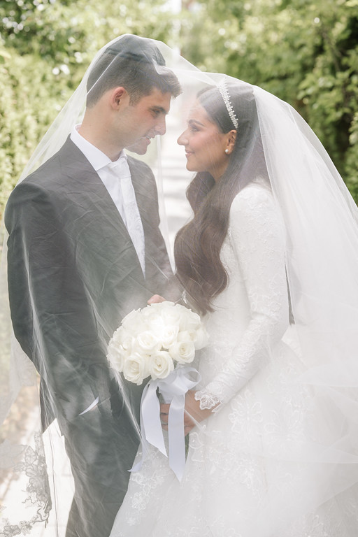 A bride and groom in wedding attire stand close under a veil, smiling at each other while leaning in and holding a bouquet of white roses outdoors.