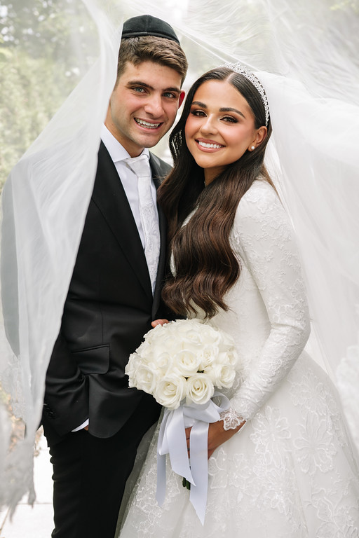 A frum wedding couple in formal attire poses under a chuppah, smiling at the camera; the bride holds a white bouquet as they stand close together.