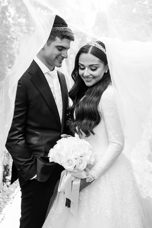 Black-and-white portrait of a bride and groom under the chuppah, smiling while the bride holds a small bouquet and the groom looks at her.