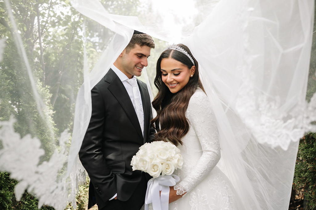 Bride in a white lace gown and headband smiles as she holds a bouquet, while the groom in a black suit stands close beside her under the chuppah canopy.