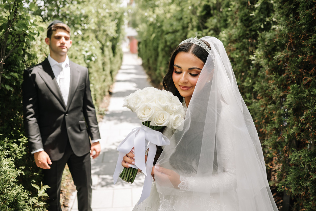 Bride in a white veil holding a bouquet of white roses outdoors while the groom stands behind her along a garden walkway.